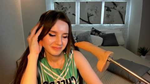 Media: Video of a young woman with long brown hair, wearing a green and white sports jersey, taking a selfie in a modern bedroom with a bed and a window with black and white abstract artwork.