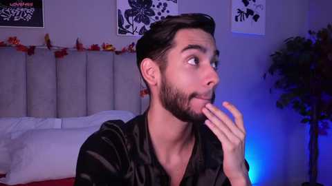 Media: Video of a surprised young man with short dark hair and a beard, wearing a black shirt, in a bedroom with white bedding, floral wall art, and a blue-lit potted plant.