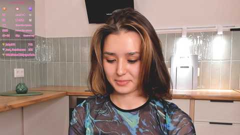 Media: Video of a young woman with shoulder-length brown hair, wearing a translucent blue and green dress, standing in a modern kitchen with white cabinets, tiled backsplash, and a refrigerator.