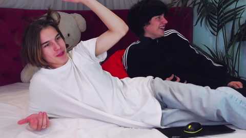 Media: Video of two young people on a bed; one with light brown hair, white shirt, and jeans, the other with dark hair, black jacket, and light pants.