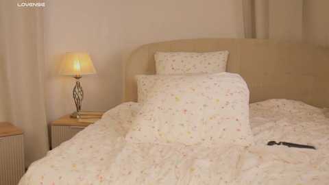 Media: Video of a cozy bedroom with a cream-colored upholstered headboard, floral bedding, a wooden nightstand, and a table lamp with a beige shade. The room exudes a warm, inviting atmosphere.