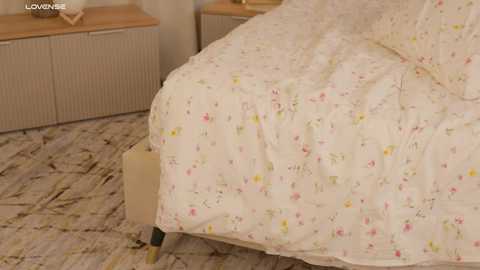 Media: A video of a neatly made bed with white floral-patterned sheets, beige wooden headboard, and light wooden floor. Background includes a beige cabinet and a patterned carpet.