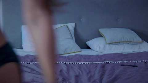 Media: A video of a dimly lit bedroom showing a neatly made bed with white pillows and a lavender comforter. The background features a gray upholstered headboard. The image is slightly blurred, focusing on the bed.