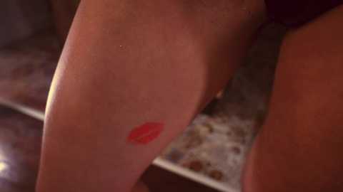 Media: A close-up video of a woman's smooth, light-skinned thigh with a prominent red lipstick kiss mark. The background shows a blurred, dark surface, possibly a table or countertop. The image focuses on the intimate and sensual nature of the kiss.