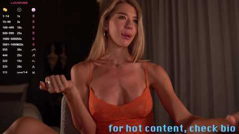 Media: Video of a blonde woman in a low-cut orange top, holding a phone, in a dimly lit room with a TV screen displaying stats. Text at the bottom reads, \"for hot content, check big.\