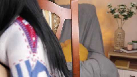 Media: Video of a woman with long black hair wearing a white blouse with red and blue embroidery, sitting in a wooden chair next to a yellow cushioned chair, in a warmly lit room with a potted plant on a shelf.