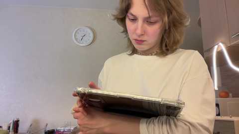 Media: Video of a young, fair-skinned girl with shoulder-length brown hair, wearing a beige sweater, intently examining a sealed brownie in a plastic container in a simple kitchen with beige walls, a clock, and utensils.