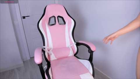 Media: A video of a pink gaming chair with white accents and black armrests, positioned in a minimalist room with light gray walls and a white door, featuring a person's arm reaching out.