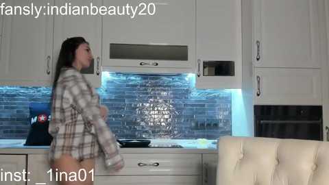 Media: Video of a young woman with long black hair, wearing a plaid shirt, dancing in a modern kitchen with white cabinets and a tiled backsplash.