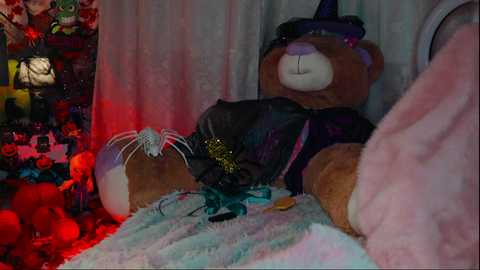 Media: Video of a plush bear in a dark, witch-themed costume lying on a soft, pink blanket. Background features a cluttered, Halloween-themed room with hanging decorations, including a green witch hat and a spider web.