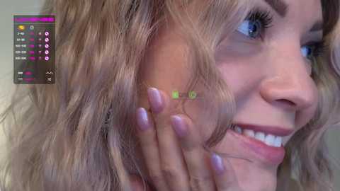 Media: Close-up video of a smiling woman with wavy, light brown hair, showcasing her blue eyes and soft pink lips. Her hand, with neatly manicured nails, gently touches her cheek. The background is blurred, with a virtual reality app interface visible on the left.