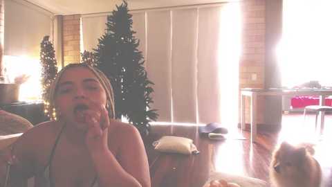 Media: Video of a young woman with braided hair, wearing a spaghetti strap top, holding a phone to her ear, sitting in a living room with a Christmas tree, sunlit room.