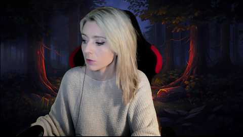 Media: Video of a young woman with straight, blonde hair, wearing a beige sweater, sitting on a black chair with red accents in a dimly lit, forest-themed background.