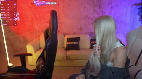 Media: Video of a dimly lit living room with red and blue lighting, featuring a blonde woman in a white top and black pants sitting in a black gaming chair, watching a game on a large screen.