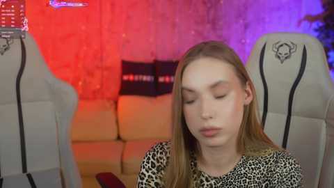 Media: Video of a young, fair-skinned woman with long, straight blonde hair, wearing a leopard-print top, seated in a gaming chair with skull decals. The background features a red and purple lit room with a couch and black pillows.