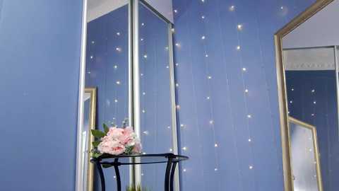 Media: A video of a cozy bedroom corner with a blue wall adorned with fairy lights and a large mirror. A small black metal table holds a bouquet of pink peonies and greenery.