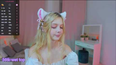 Media: Video of a blonde woman with light skin, wearing cat ears, pink eyeshadow, and a white off-shoulder top, smiling in a pink-lit room with a vanity table.