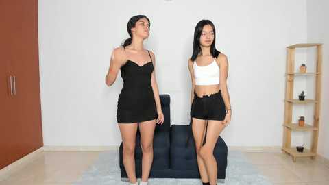 Media: Video of two young women with medium skin tones, one in a black mini dress, the other in a white crop top and shorts, standing in a minimalist living room with white walls, a blue chair, and wooden shelves.