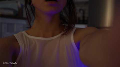 Media: A dimly lit video of a young woman with light skin and shoulder-length brown hair, wearing a sheer white tank top, capturing a close-up view of her chest and upper body.