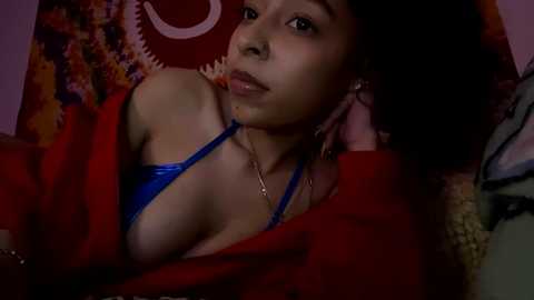 Media: A dimly lit video of a young African-American woman with a medium-dark complexion, wearing a blue bra, lying on a red bedspread, surrounded by colorful patterns.