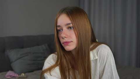 Media: Video of a young woman with long, straight, auburn hair, wearing a white shirt, sitting on a bed with gray pillows, in a dimly lit room with gray curtains.