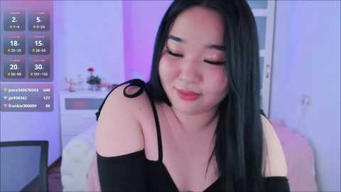 Media: Video of an East Asian woman with long black hair, wearing a black off-shoulder top, smiling, in a softly lit bedroom.