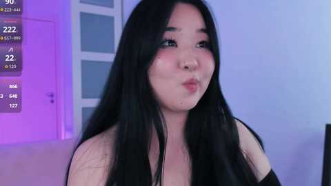 Media: Video of an East Asian woman with long black hair, fair skin, and a full figure, wearing a black top, indoors with purple lighting, and a digital screen showing numbers in the background.