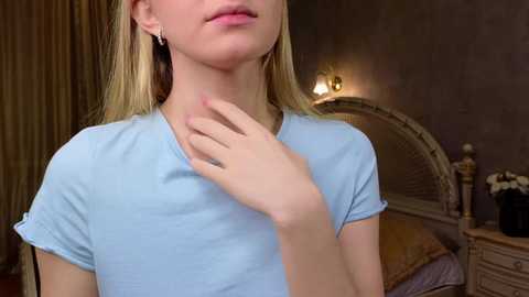 Media: Video of a young blonde woman with light skin, wearing a light blue shirt, touching her neck in a dimly lit bedroom with a vintage bed and wooden furniture.