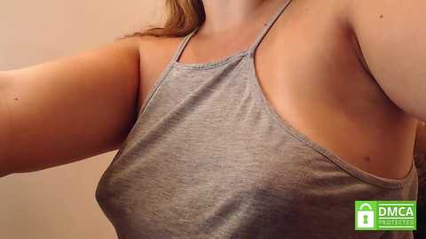 Media: A video of a fair-skinned woman with a slender physique wearing a gray tank top, showcasing her bare shoulder and a mole on her upper arm. The background is a neutral, beige color.