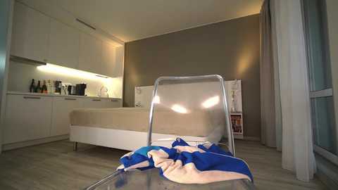 Media: Video of a modern, minimalist bedroom with beige walls, white cabinets, a bed, and a clear chair holding a blue and white towel.