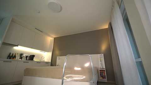 Media: A modern, minimalist kitchen with beige cabinets, stainless steel appliances, a clear acrylic chair, and a white fridge against a taupe wall.