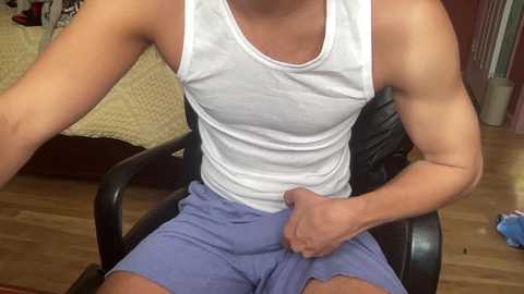 Media: Video of a muscular man with light skin in a white tank top and blue shorts, sitting on a black chair. Background shows a bed with beige sheets and a wooden floor.