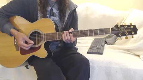 Media: Video of a person with long, curly hair playing a wooden acoustic guitar on a white couch. They wear a dark jacket over a blue graphic tee. A black keyboard is placed beside them.