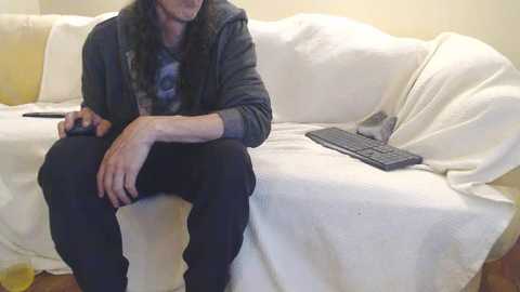 Media: Video of a man with long hair, wearing a dark hoodie and jeans, sitting on a white couch, holding a remote, with a keyboard and laptop nearby.