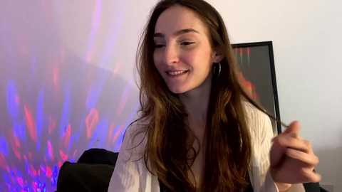 Media: A video of a young woman with long brown hair, smiling, wearing a white blouse, in a brightly lit room with colorful LED lights and a black TV in the background.