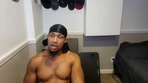 Media: Video of a shirtless, muscular Black man with a black baseball cap, sitting on a black exercise bench in a dimly lit, sparsely furnished room with white walls and a few hanging shoes.