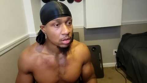 Media: Video of a shirtless, muscular Black man with a black headband and beard, posing in a room with white cabinets, a black duffel bag, and a workout mat on the beige carpeted floor.