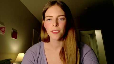 Media: Video of a young Caucasian woman with straight, shoulder-length light brown hair, wearing a lavender cardigan, in a dimly lit room with beige walls, a door, and a lamp.