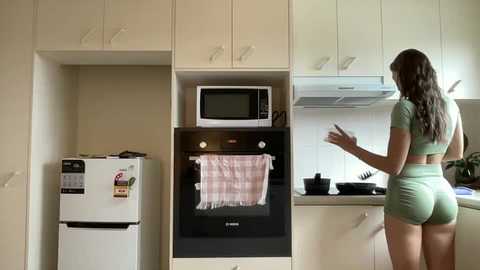 Media: Video of a modern kitchen with white cabinets and a microwave oven. A woman with long brown hair, wearing a green crop top and shorts, stands at the counter, back to the camera.