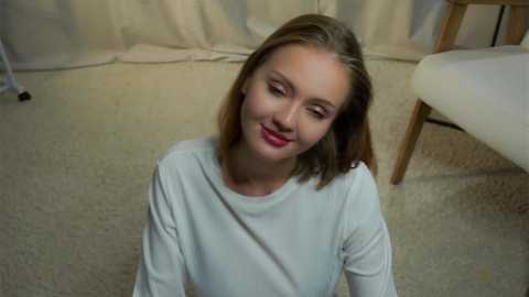 Media: Video of a young Caucasian woman with fair skin, straight brown hair, and a slight smile, wearing a white long-sleeve shirt, sitting on beige carpet in a softly lit room with cream curtains and a wooden chair.