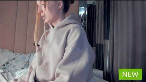 Media: Video of a fair-skinned woman in a beige hoodie, sitting on a bed with floral-patterned sheets in a dimly lit room.