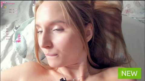 Media: A video of a young woman with fair skin, light brown hair, and closed eyes lying on a bed with a patterned white and pastel-colored pillowcase, wearing a necklace.