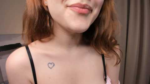 Media: Video of a young woman with fair skin and reddish-brown wavy hair, wearing a black strapless top. She has a small heart tattoo on her right shoulder. Background shows a beige curtain and part of a bed.