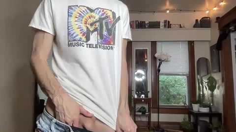 Media: Video of a white man in a white \"Music Television\" t-shirt, slightly lifting his shirt to expose his bare stomach, standing in a cozy living room with a window and plant.