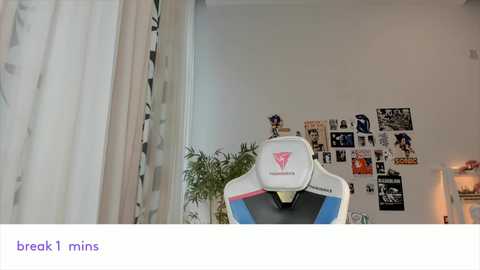 Media: Video of a modern room with a white, minimalist couch adorned with pink and blue pillows. A white headset sits atop the couch, partially visible. The background features a white wall with a collage of black-and-white photos and a potted plant.