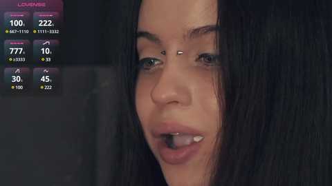 Media: A close-up video of a young woman with long, straight black hair, light brown skin, and slightly parted lips, showing a small black object. The background is dark, with a digital health monitoring display on the left.