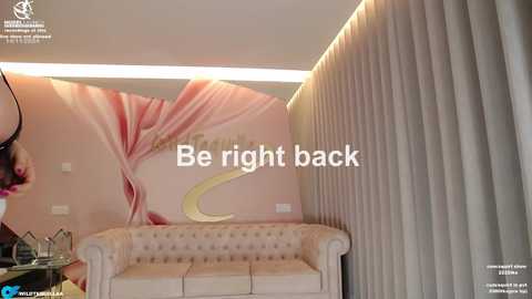 Media: Video of a luxurious spa interior featuring a beige tufted sofa, pink and white abstract wall mural, and vertical blinds. Text overlay: \"Be right back.\