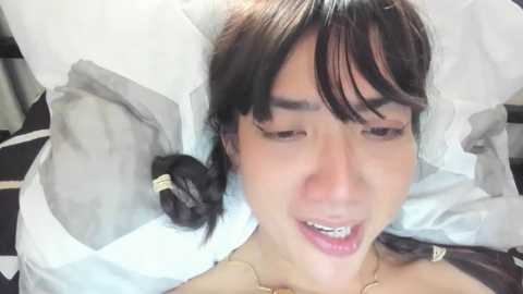 Media: A close-up video of a young Asian woman with fair skin, brown hair, and slightly parted lips, lying on a white pillow, wearing a beige top.