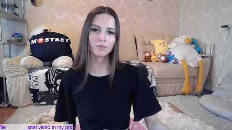 Media: A young Caucasian woman with long brown hair, fair skin, and a slender build, wearing a black top, sits in a cozy living room with plush toys and a beige sofa.