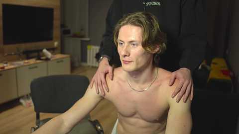 Media: Video of a shirtless, fair-skinned, muscular young man with shoulder-length brown hair, receiving a massage from a person wearing a black shirt. Background includes a TV, wooden shelves, and a dark sofa.